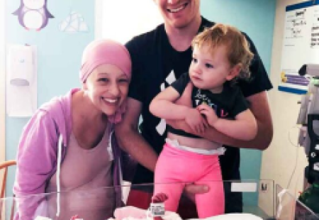 A breast cancer patient and mother smiles with her husband and toddler next to their newborn in the NICU  